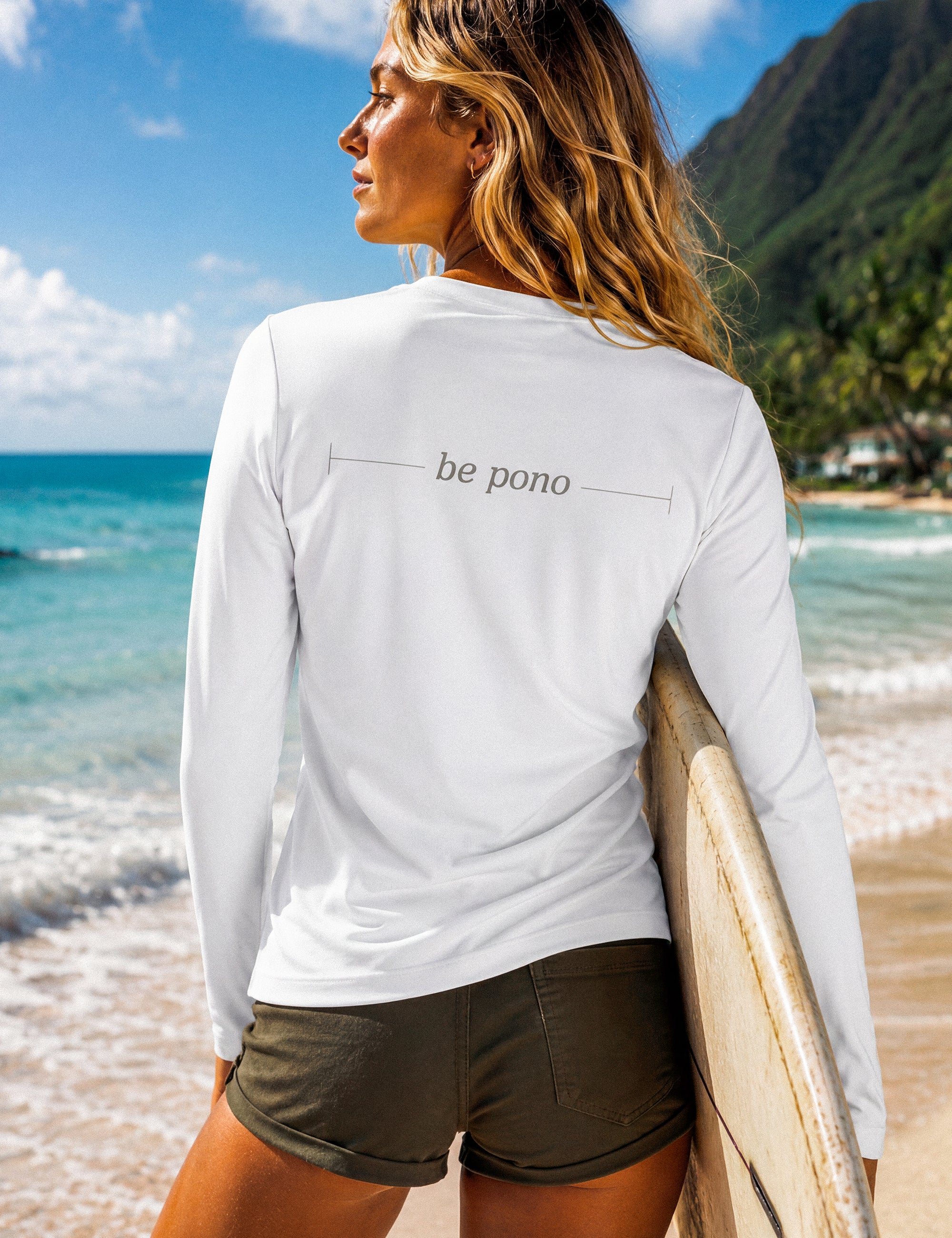 Woman holding a surfboard on a beach with 'be pono' shirt