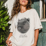 woman wearing a bone colored t-shirt with a puffer fish graphic standing in front of a wood house