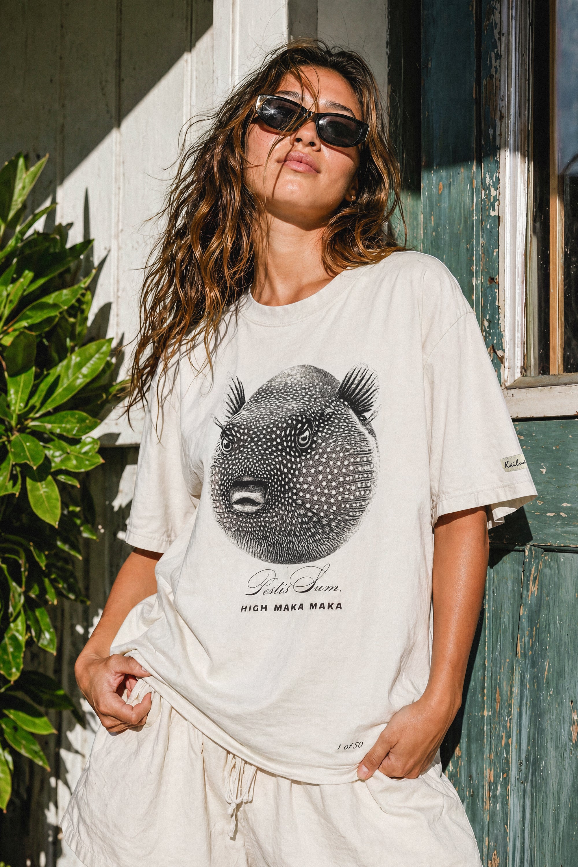 woman wearing a bone colored t-shirt with a puffer fish graphic standing in front of a wood house