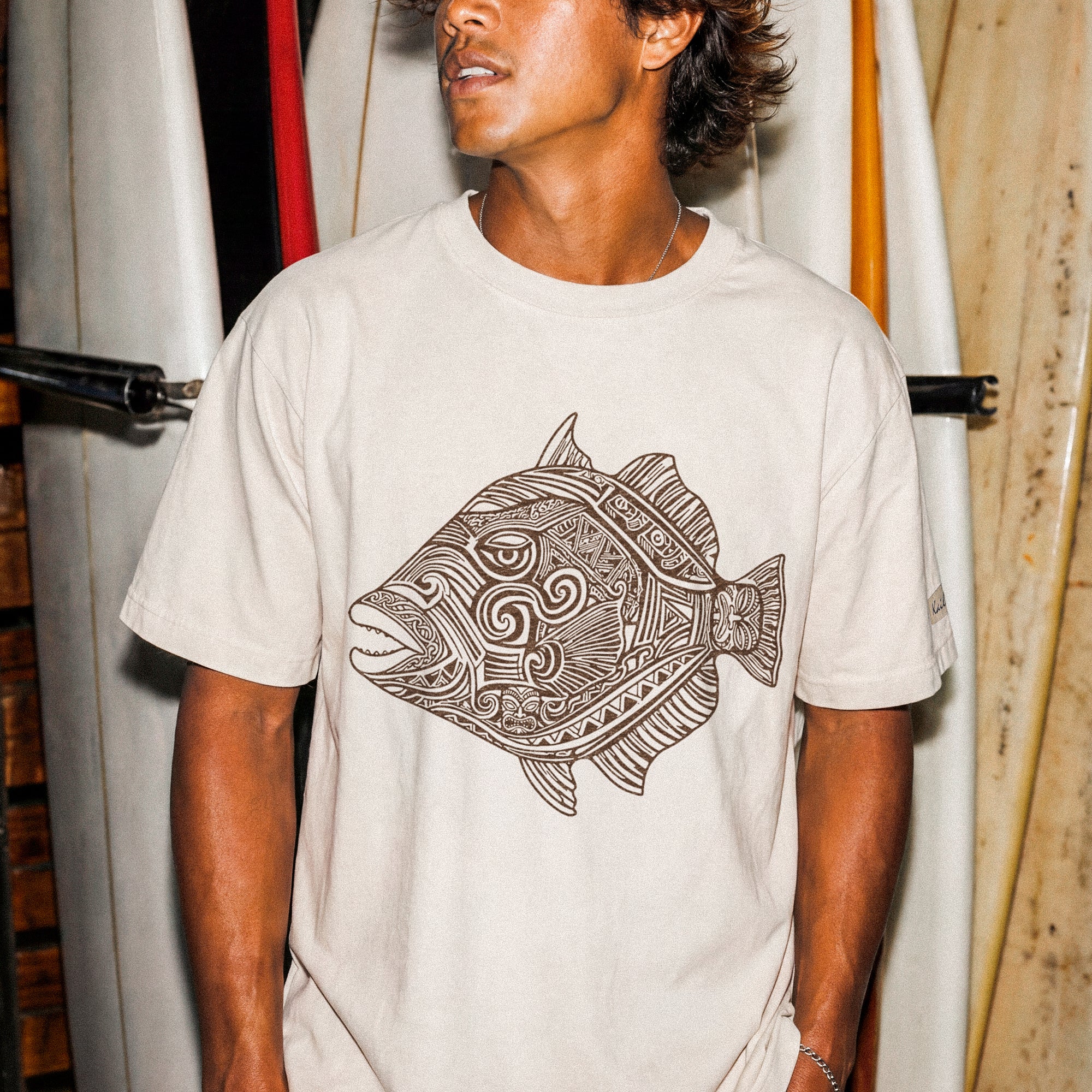 kailua hi surf tees humuhumu men's luxe tee in pearl brown – three-quarter lifestyle shot of a male model looking away, standing in front of a vertical rack of stacked white surfboards. he is wearing a relaxed pearl-colored graphic shirt featuring a large brown intricate tribal woodcut style illustration of a humuhumu fish with swirling geometric patterns, paired with dark shorts