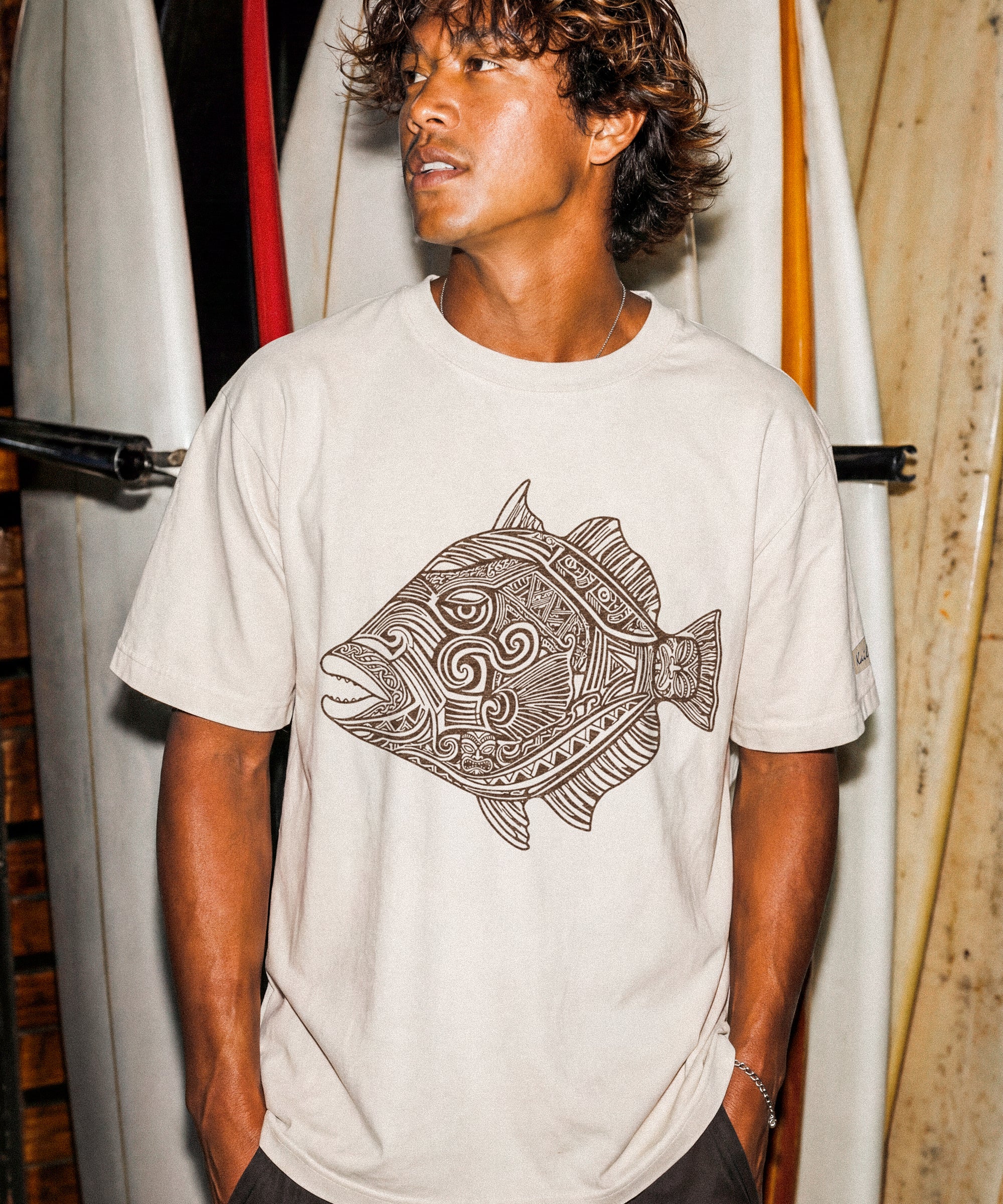kailua hi surf tees humuhumu men's luxe tee in pearl brown – three-quarter lifestyle shot of a male model looking away, standing in front of a vertical rack of stacked white surfboards. he is wearing a relaxed pearl-colored graphic shirt featuring a large brown intricate tribal woodcut style illustration of a humuhumu fish with swirling geometric patterns, paired with dark shorts