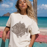 kailua hi surf tees humuhumu women's luxe tee in pearl brown – close-up lifestyle shot of a blonde female model leaning against a colorful pink and teal painted stone wall. she is wearing a relaxed pearl-colored graphic shirt featuring a large brown intricate tribal woodcut style humuhumu fish illustration, paired with light trousers, against a turquoise ocean, island, and palm tree background