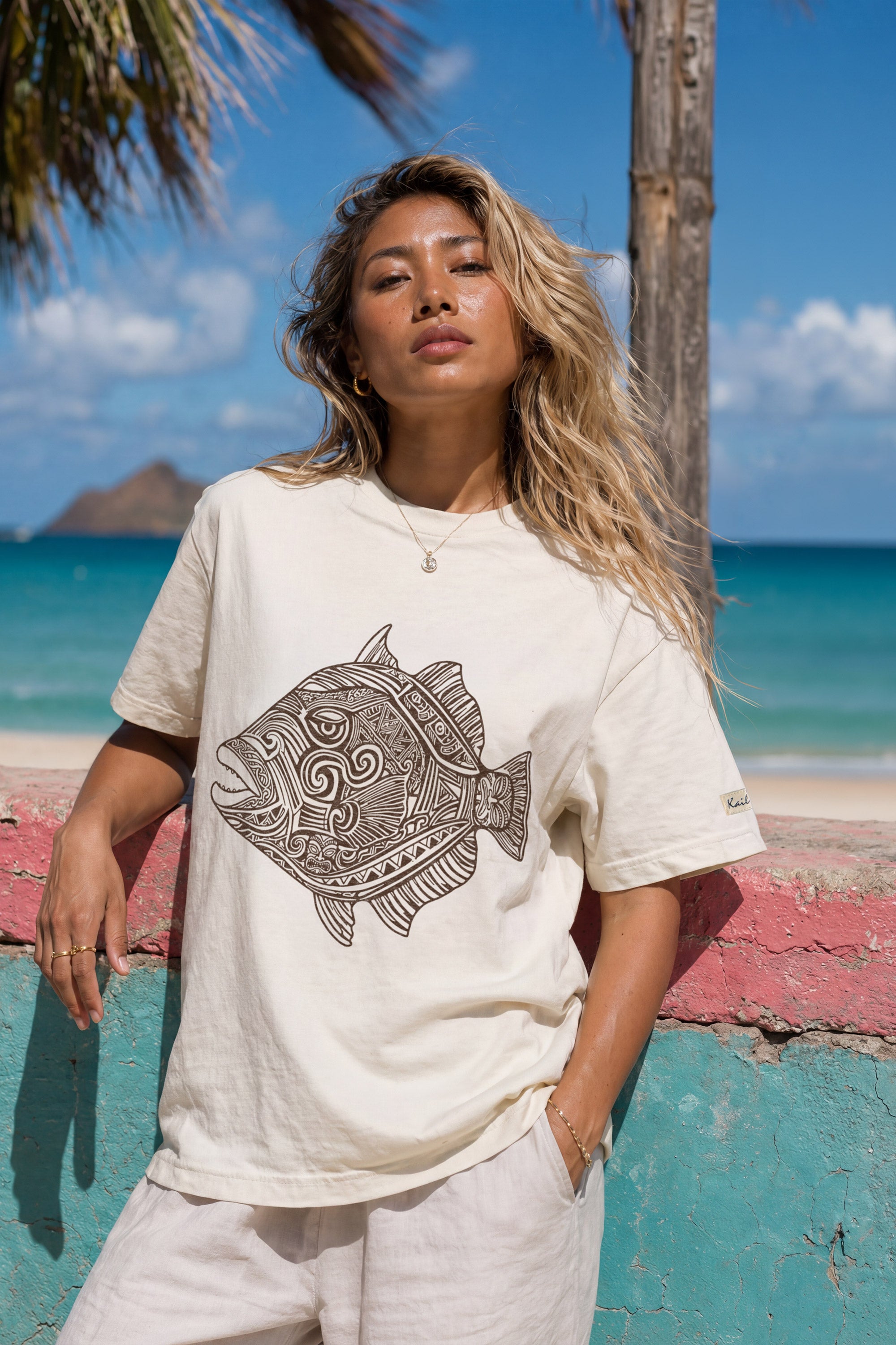 kailua hi surf tees humuhumu women's luxe tee in pearl brown – close-up lifestyle shot of a blonde female model leaning against a colorful pink and teal painted stone wall. she is wearing a relaxed pearl-colored graphic shirt featuring a large brown intricate tribal woodcut style humuhumu fish illustration, paired with light trousers, against a turquoise ocean, island, and palm tree background