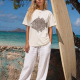 kailua hi surf tees humuhumu women's luxe tee in pearl brown – full-length lifestyle shot of a blonde female model standing barefoot on a sandy beach next to a large vertical wooden surfboard. she is wearing a relaxed pearl-colored graphic shirt featuring a large brown intricate tribal woodcut style humuhumu fish illustration, paired with loose white linen trousers, against a turquoise ocean and island backdrop