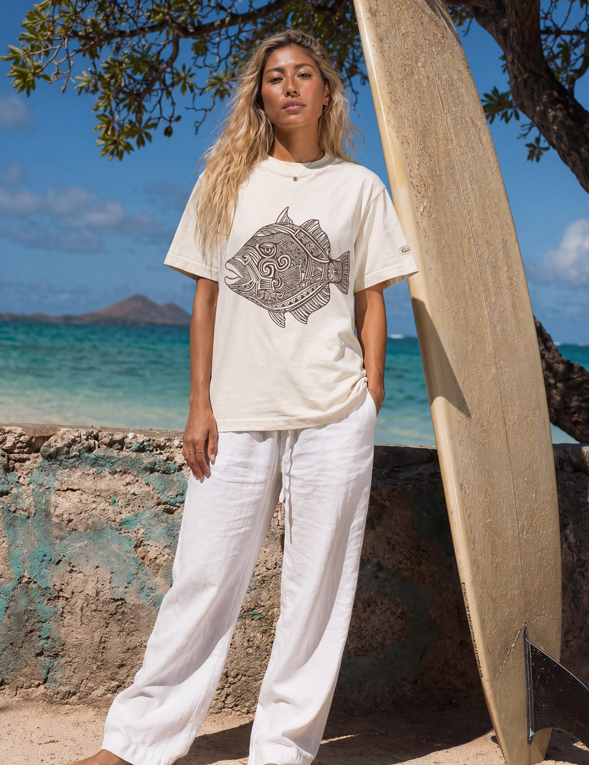kailua hi surf tees humuhumu women's luxe tee in pearl brown – full-length lifestyle shot of a blonde female model standing barefoot on a sandy beach next to a large vertical wooden surfboard. she is wearing a relaxed pearl-colored graphic shirt featuring a large brown intricate tribal woodcut style humuhumu fish illustration, paired with loose white linen trousers, against a turquoise ocean and island backdrop