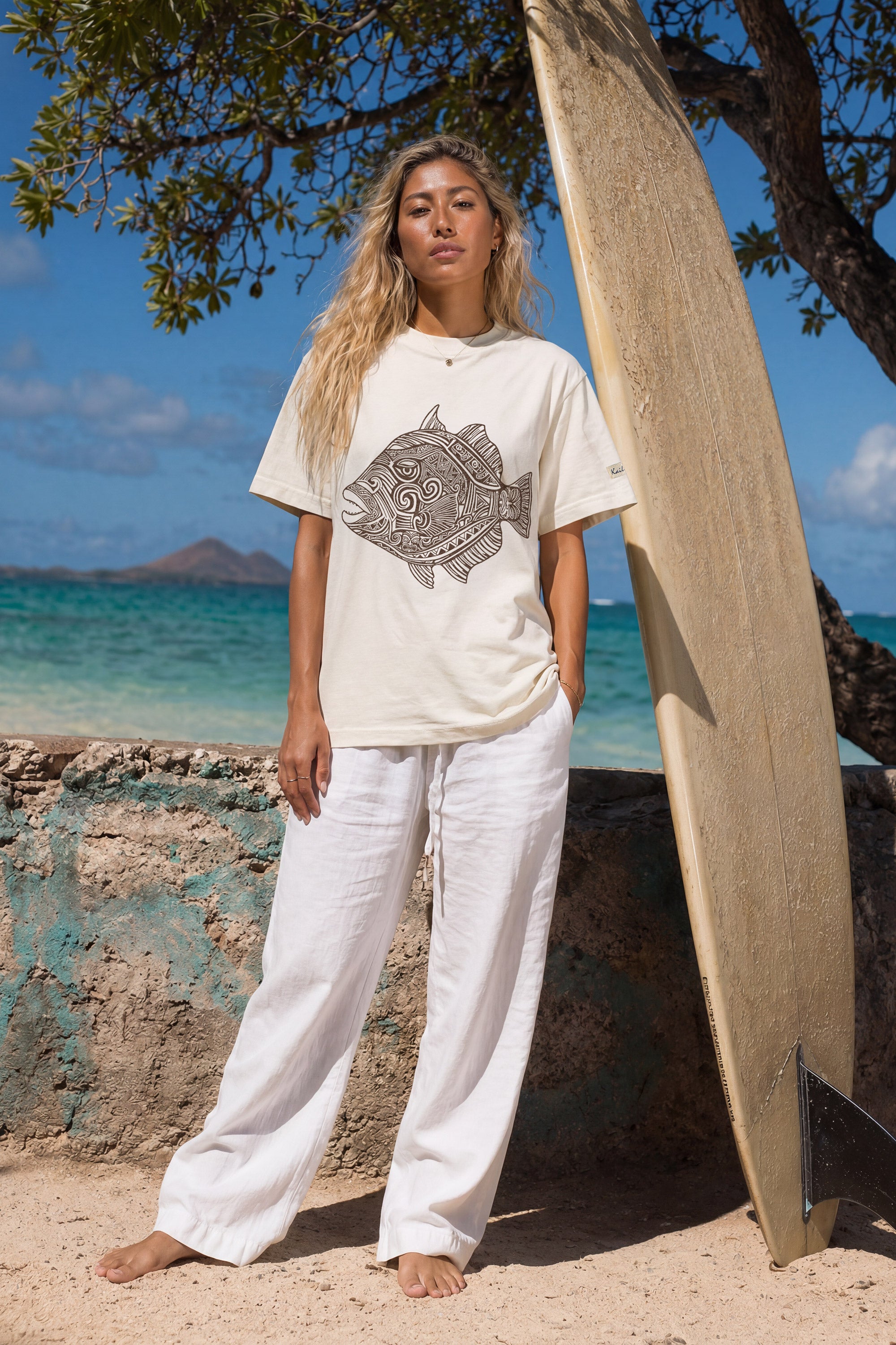kailua hi surf tees humuhumu women's luxe tee in pearl brown – full-length lifestyle shot of a blonde female model standing barefoot on a sandy beach next to a large vertical wooden surfboard. she is wearing a relaxed pearl-colored graphic shirt featuring a large brown intricate tribal woodcut style humuhumu fish illustration, paired with loose white linen trousers, against a turquoise ocean and island backdrop