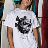 kailua hi surf tees pilau premium tee in white – waist-up men's lifestyle shot of a model standing in front of a rack of stacked surfboards. he is wearing a silver chain necklace and a relaxed white graphic shirt featuring a spiky black and white porcupine fish design with a spiral eye, cursive putridus text, and loose black trousers