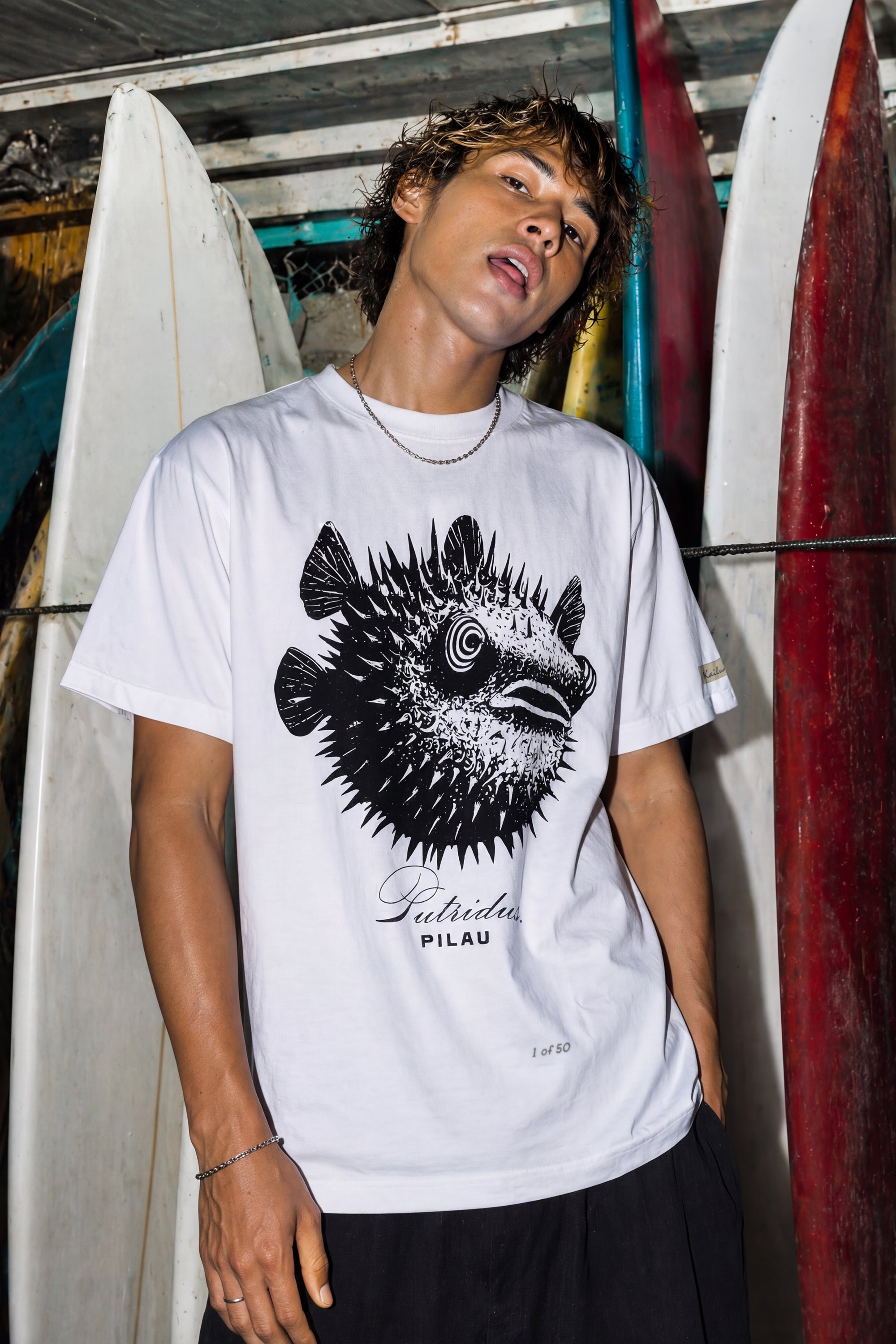 kailua hi surf tees pilau premium tee in white – waist-up men's lifestyle shot of a model standing in front of a rack of stacked surfboards. he is wearing a silver chain necklace and a relaxed white graphic shirt featuring a spiky black and white porcupine fish design with a spiral eye, cursive putridus text, and loose black trousers