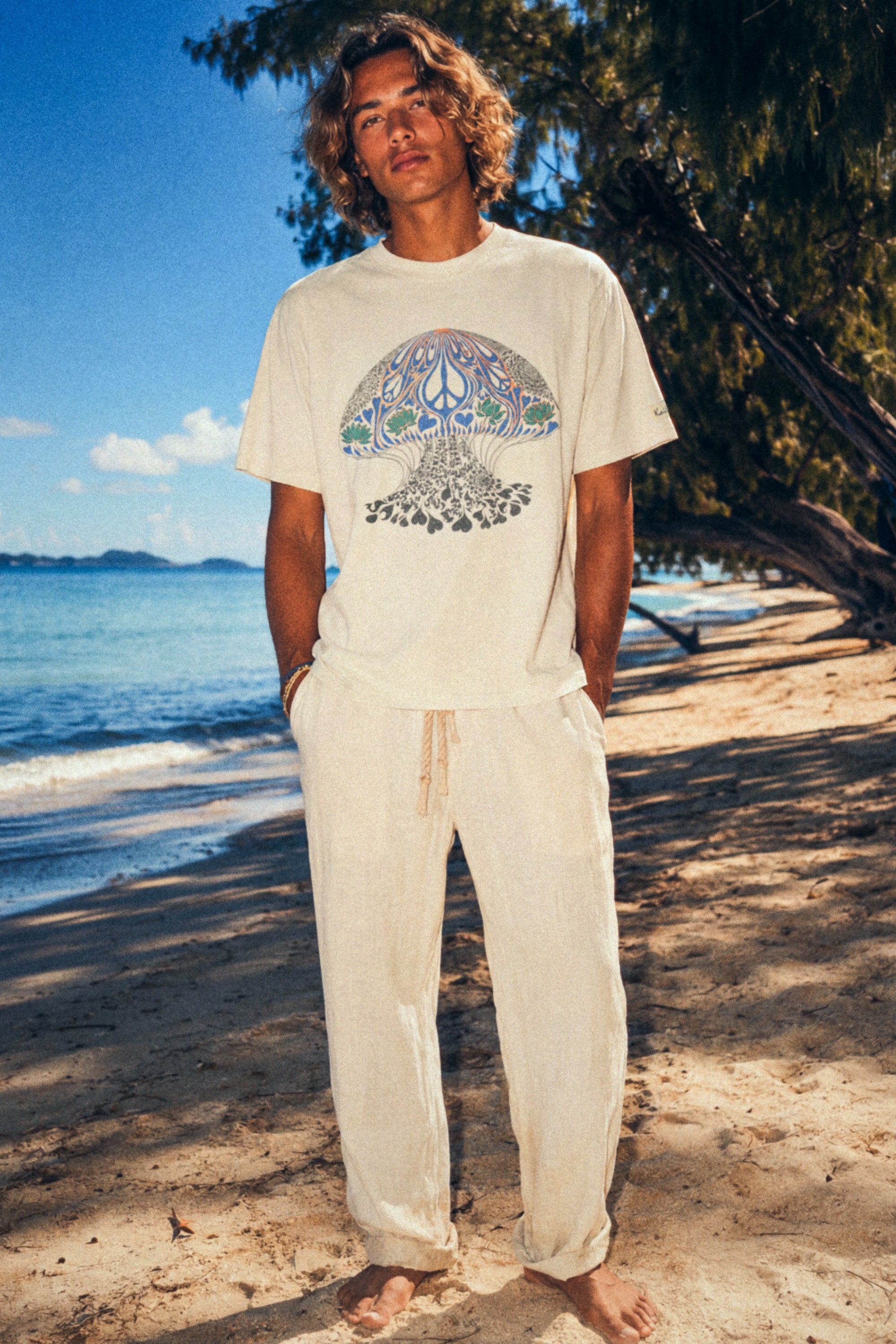 kailua hi surf tees psychedelic moon jelly luxe men's tee in faded ecru – full-body beach lifestyle shot of a model standing barefoot on the sand wearing the relaxed fit graphic shirt paired with light linen drawstring trousers
