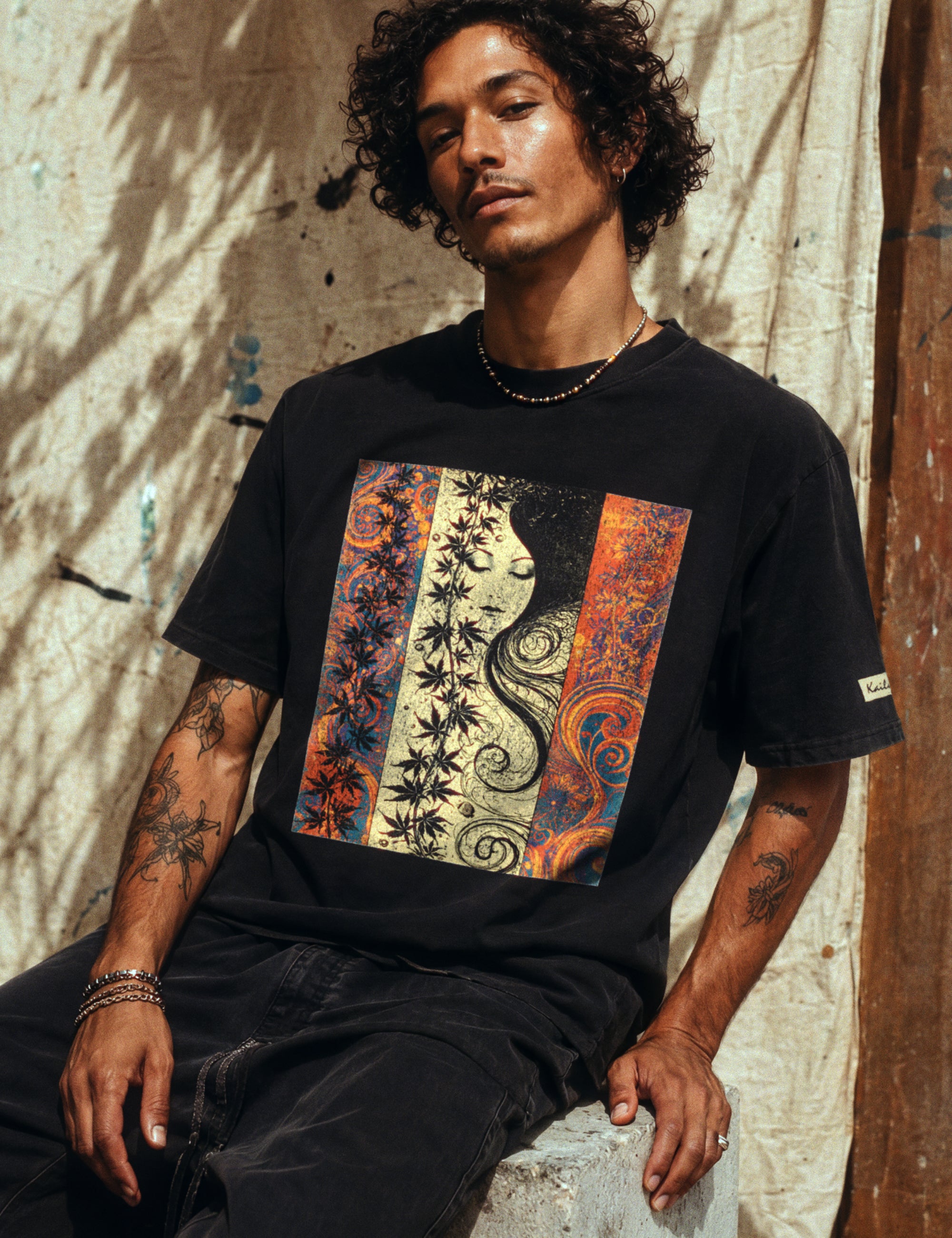 kailua hi surf tees seaweed pua o ke kai men's luxe tee in faded black – three-quarter lifestyle shot of a male model with curly hair and arm tattoos sitting on a concrete block against a sunlit canvas backdrop. he wears a relaxed faded black graphic shirt featuring a colorful art nouveau portrait panel with a woman's face, stylized botanical leaves, and orange and blue swirls, paired with dark trousers
