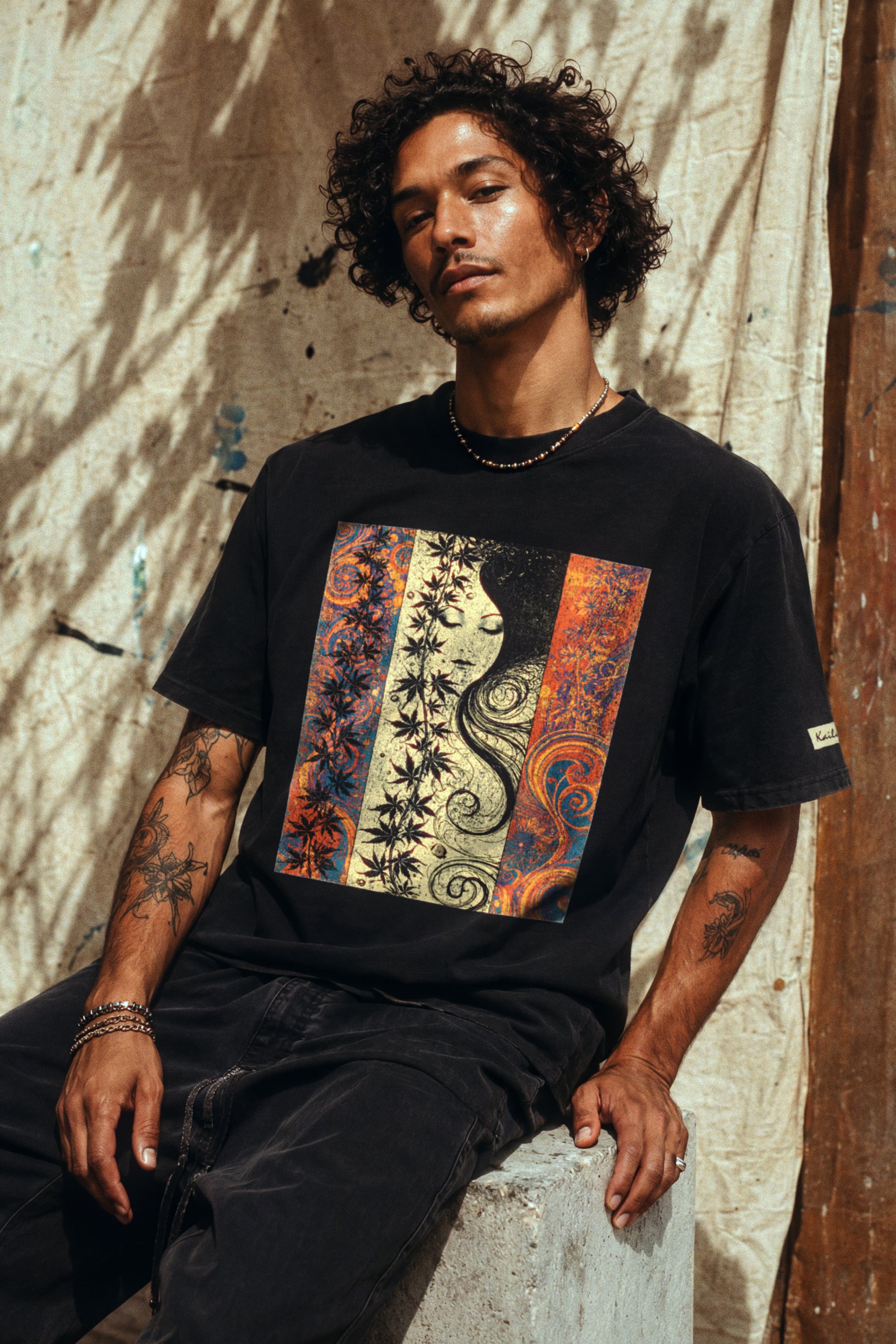 kailua hi surf tees seaweed pua o ke kai men's luxe tee in faded black – three-quarter lifestyle shot of a male model with curly hair and arm tattoos sitting on a concrete block against a sunlit canvas backdrop. he wears a relaxed faded black graphic shirt featuring a colorful art nouveau portrait panel with a woman's face, stylized botanical leaves, and orange and blue swirls, paired with dark trousers
