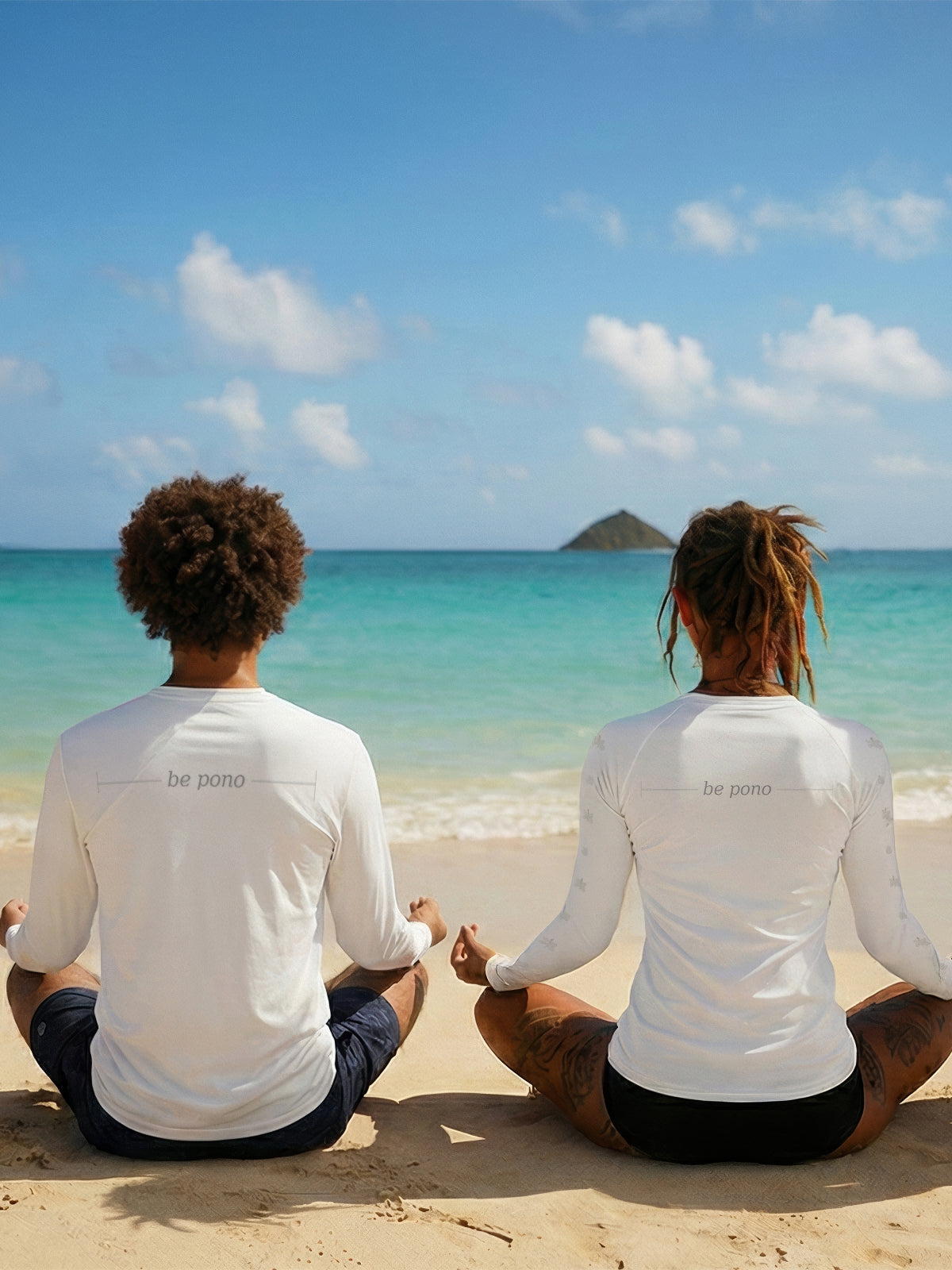 Two people sitting on a beach wearing white shirts with text, facing the ocean.